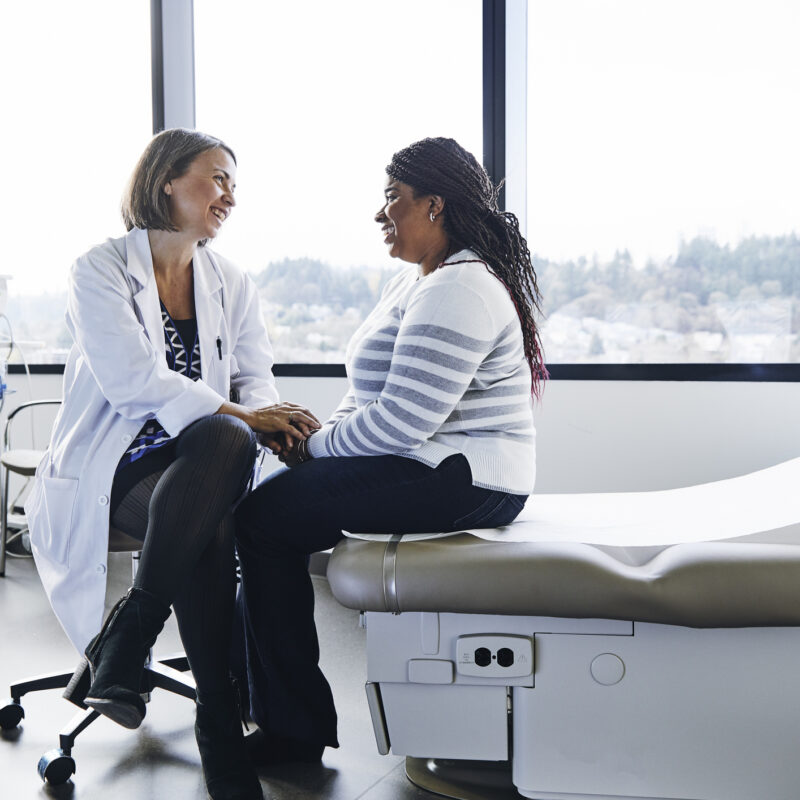 A white woman doctor speaking with a woman patient in an exam room. The patient is a middle-age Black woman who is clothed in blue jeans and a white sweater. Both women are smiling.