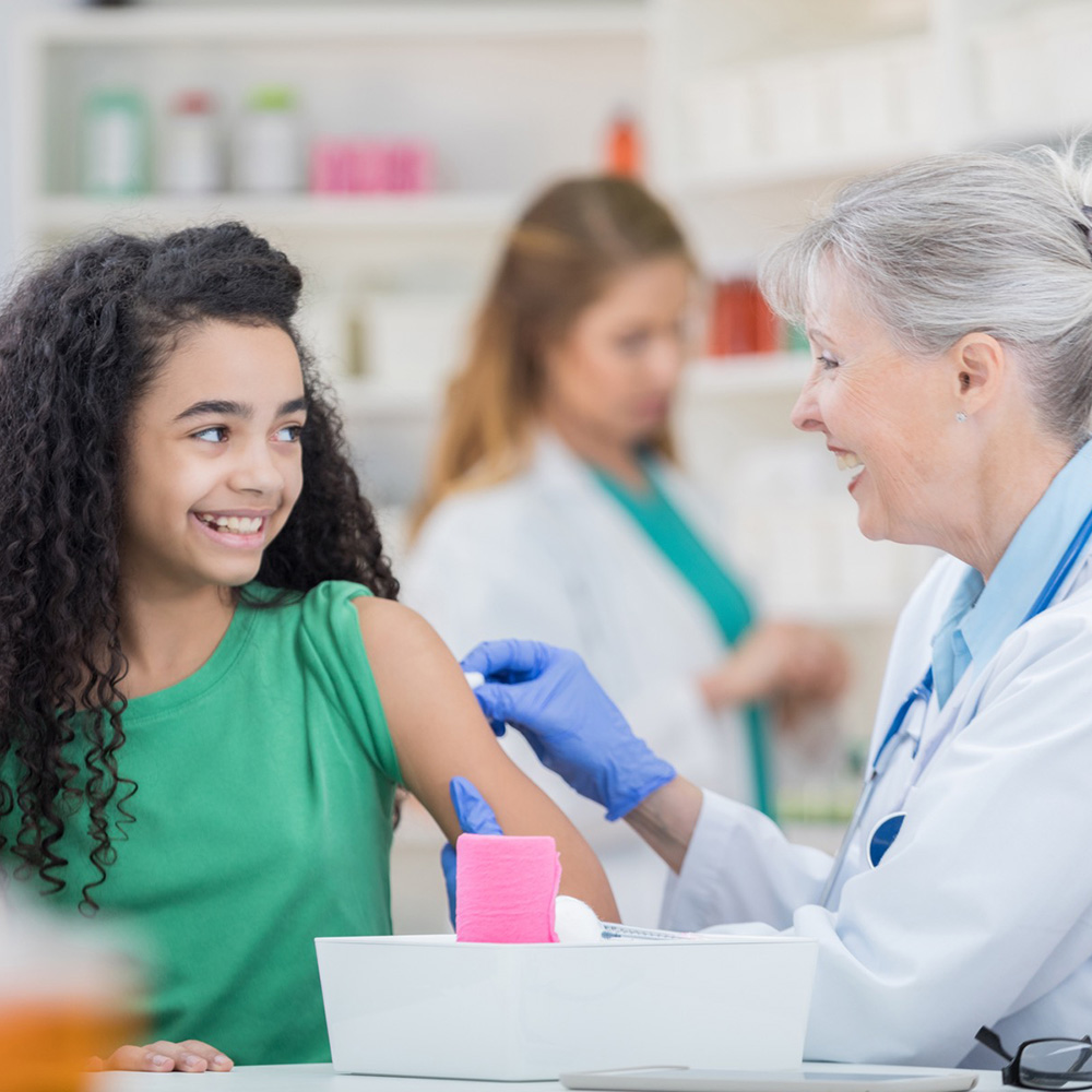 A girl with brown skin and long, dark curly hair is preparing to receive a shot from a woman pharmacist.